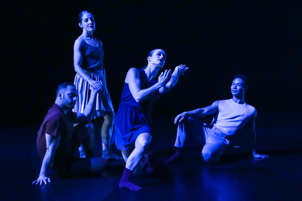 a tableau of dancers bathed in blue light, a woman, the central figure a woman leans to one side, and the figures around her stare at her thoughtfully
